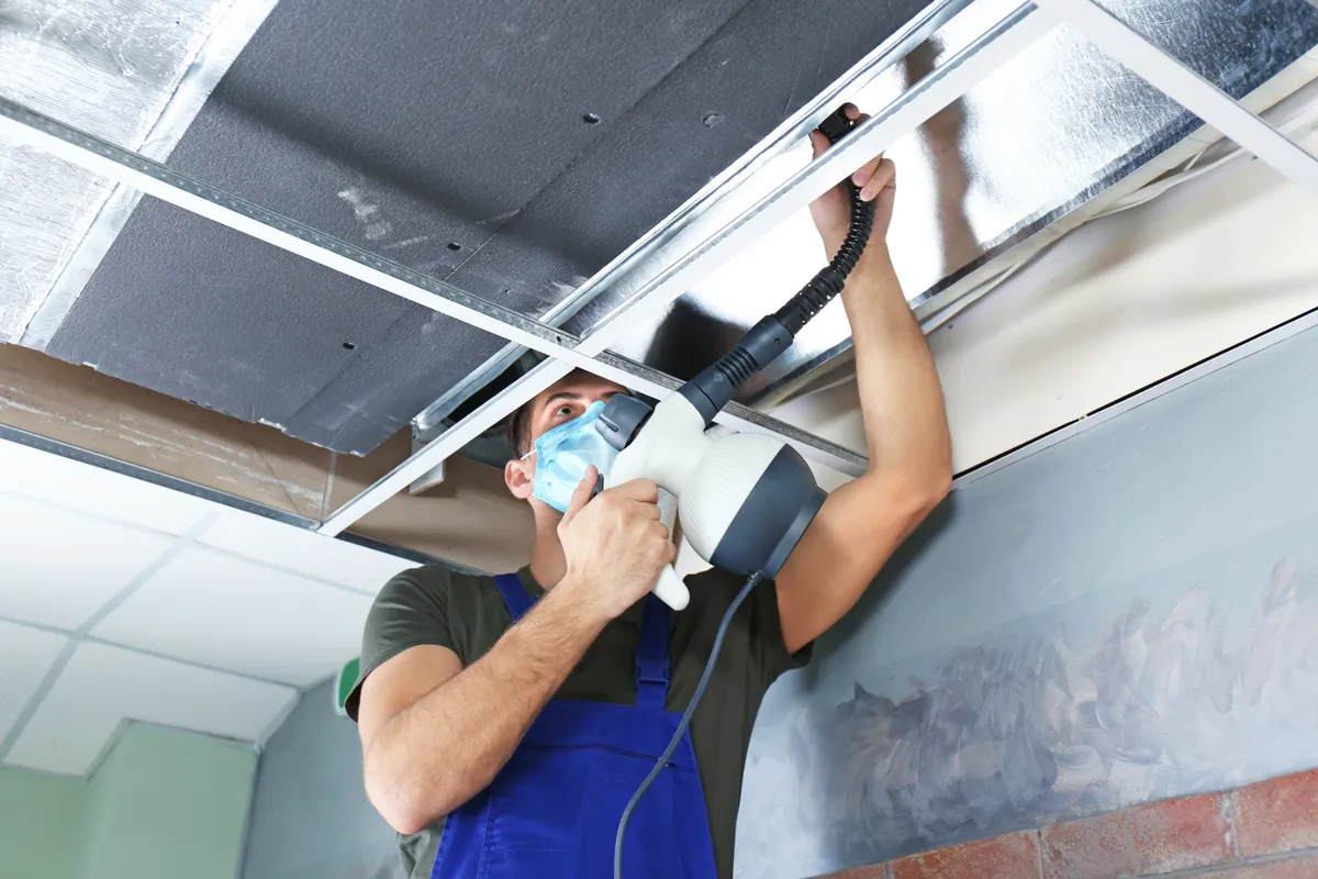Worker in a mask cleaning a ceiling air duct with a handheld tool