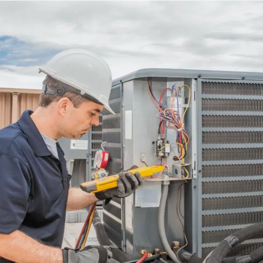 HVAC technician in a hard hat testing an outdoor air conditioner with a multimeter